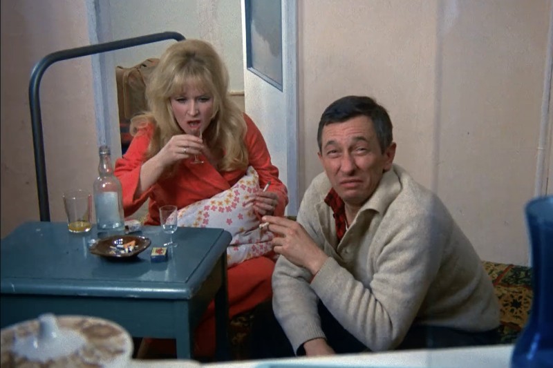 Kobieta i mężczyzna siedzą na podłodze przy małym stoliku z napojami i popielniczką. Kobieta trzyma szklankę i wygląda na zaskoczoną, podczas gdy mężczyzna krzywi się, pijąc. Sceneria wydaje się swobodna i domowa.