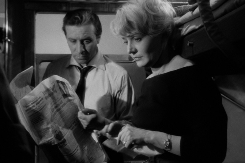 Mężczyzna w koszuli i krawacie czyta gazetę, a obok niego stoi kobieta o krótkich blond włosach. Oboje wyglądają poważnie, a wszystko to w środku, który wygląda jak przedział pociągu.