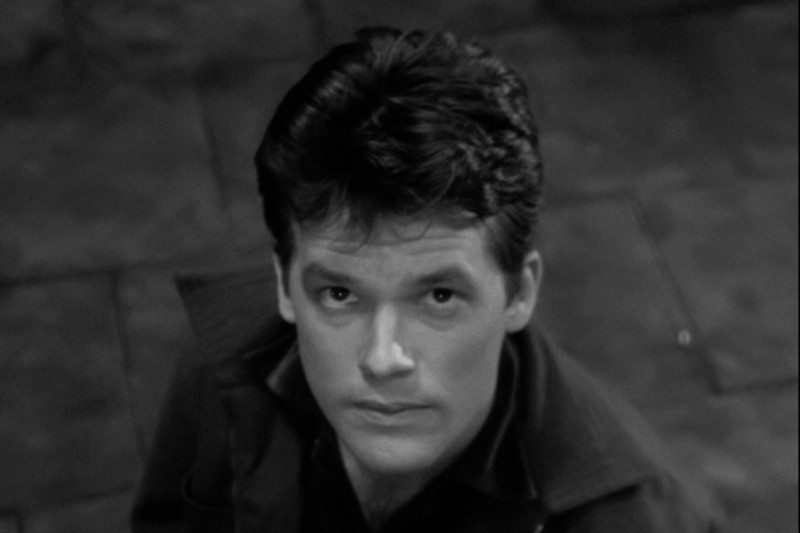 Młody mężczyzna o ciemnych włosach i poważnym wyrazie twarzy spogląda w górę w stronę kamery. Obraz jest czarno-biały, a tło przedstawia kafelkową lub kamienną podłogę.