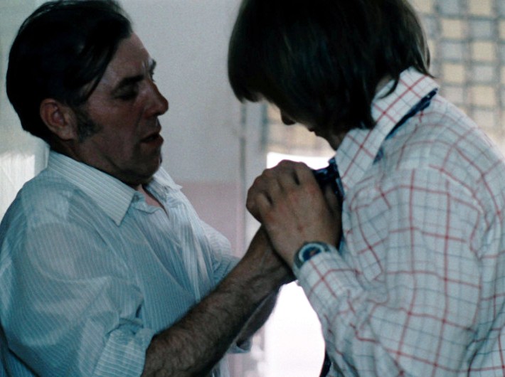 Dwóch mężczyzn stoi blisko siebie w pomieszczeniu; jeden w białej koszuli pomaga drugiemu, który ma na sobie kraciastą koszulę i krawat, poprawić krawat. Głowa drugiego mężczyzny jest pochylona, gdy skupiają się na zadaniu. Scena wydaje się napięta i intymna.