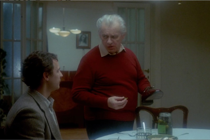 Dwóch mężczyzn znajduje się w słabo oświetlonym pokoju; jeden siedzi przy stole ze słoikiem i szklanką, podczas gdy drugi, ubrany w czerwony sweter, stoi obok niego, trzymając portfel. Zdjęcie wisi na ścianie w tle.