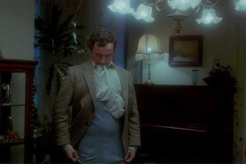 Mężczyzna w garniturze stoi w słabo oświetlonym pokoju z koszulą naciągniętą na połowę głowy i rękami w niebieskich spodniach. W pokoju znajdują się zabytkowe meble, lampa, żyrandol i rośliny.