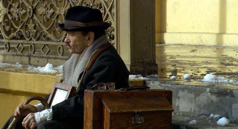 Starsza osoba w płaszczu, szaliku i czapce siedzi na ławce w pobliżu zaśnieżonego, ozdobnego budynku, trzyma ramkę ze zdjęciem i laskę, obok niej leży drewniane pudełko i okulary.