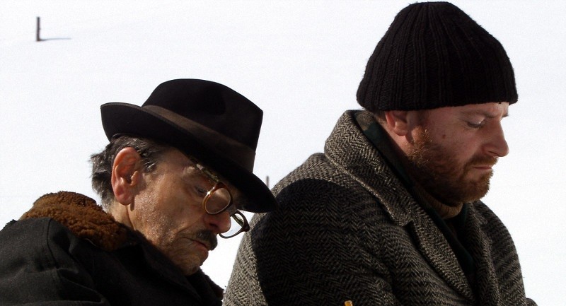 Dwóch mężczyzn w zimowych płaszczach siedzi obok siebie na śnieżnobiałym tle. Starszy mężczyzna nosi okulary i czarny kapelusz, z zamkniętymi oczami, podczas gdy młodszy mężczyzna nosi dzianinową czapkę i patrzy w dół.