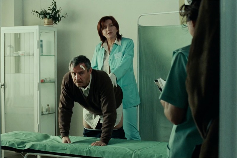 Mężczyzna ze spuszczonymi spodniami klęczy na szpitalnym stole do badań, podczas gdy pracownica służby zdrowia w fartuchu laboratoryjnym stoi za nim. Inna osoba w fartuchu robi notatki w pobliżu. Miejscem akcji jest gabinet badań lekarskich.