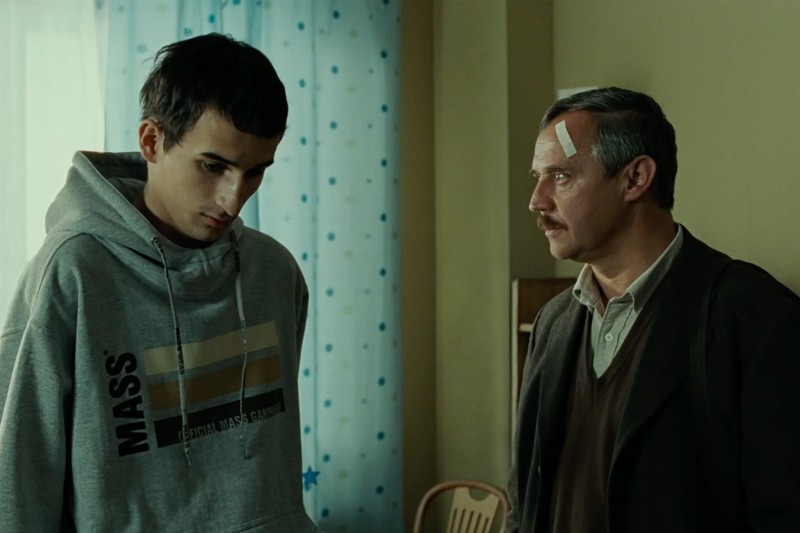 Młody mężczyzna w szarej bluzie z kapturem i bandażami na nosie stoi obok starszego mężczyzny z siwymi włosami i bandażem na czole. Są w środku, przy oknie z niebieskimi i białymi zasłonami.