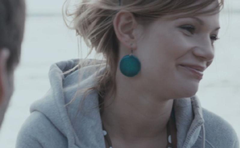 Kobieta z jasnobrązowymi włosami w nieuporządkowanym upiętym koku, ubrana w szarą bluzę z kapturem i duże turkusowe kolczyki, uśmiecha się, patrząc lekko na bok. Tło wydaje się być na zewnątrz i jest rozmyte.