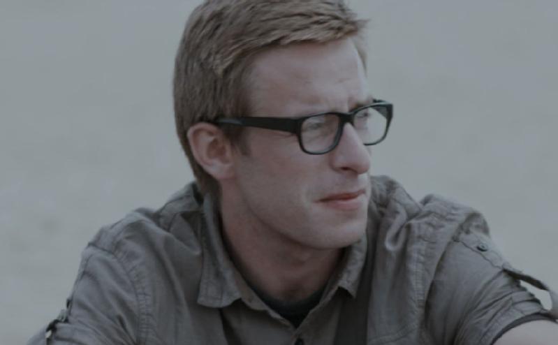 Mężczyzna z krótkimi, jasnobrązowymi włosami i czarnymi okularami patrzy w bok, ubrany w szarą koszulę z kołnierzykiem. Tło jest nieostre, wydaje się jasne i neutralne.