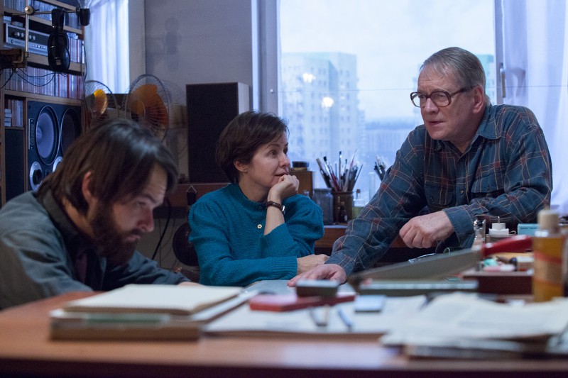 Trzy osoby siedzą i rozmawiają przy zagraconym biurku w pokoju z dużymi oknami. Mężczyzna po lewej patrzy w dół, podczas gdy kobieta i starszy mężczyzna po prawej rozmawiają i wyglądają na zamyślonych.