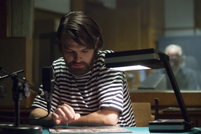 Mężczyzna z brodą i włosami do ramion, ubrany w czarno-białą koszulę w paski, siedzi przy stole pod lampą biurkową, wskazując na papier. W pobliżu znajduje się mikrofon, a inna osoba jest rozmazana w tle.