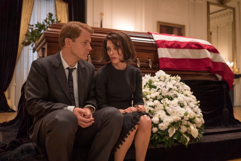 Mężczyzna i kobieta, ubrani w ciemne, formalne stroje, siedzą smutno przed trumną okrytą amerykańską flagą, obok nich leży duży, biały bukiet kwiatów, w ozdobnym, ponurym pomieszczeniu.