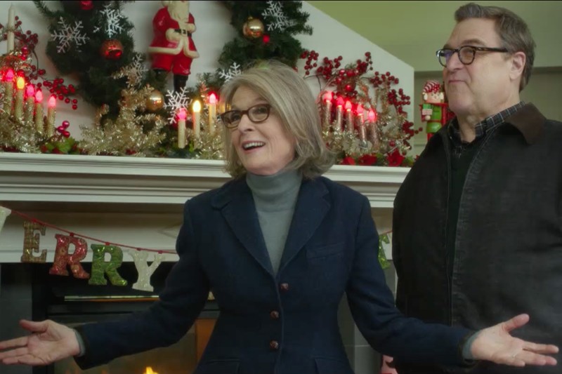 Kobieta i mężczyzna stoją przed udekorowanym kominkiem z zapalonymi świecami, świątecznym wieńcem i transparentem MERRY. Kobieta gestykuluje z otwartymi ramionami, oboje się uśmiechają w radosnym, świątecznym otoczeniu.
