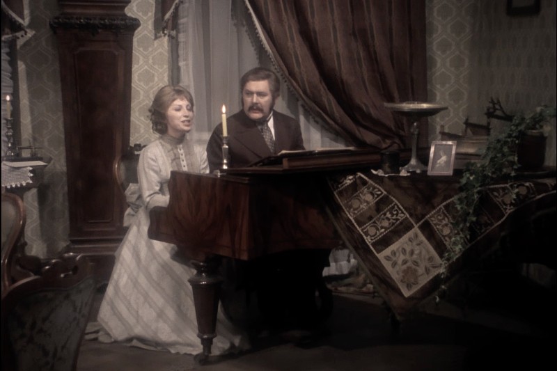 Kobieta i mężczyzna siedzą razem przy pianinie w pokoju w stylu vintage, śpiewając lub grając muzykę. Kobieta nosi jasną sukienkę, a mężczyzna garnitur i wąsy. Pokój jest udekorowany wzorzystą tapetą i ozdobnymi meblami.