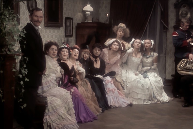 Grupa kobiet w sukienkach z epoki wiktoriańskiej siedzi w rzędzie w zabytkowym pokoju, obok nich stoi mężczyzna, a w tle muzyk gra na instrumencie dętym. Sceneria wydaje się formalna i staromodna.