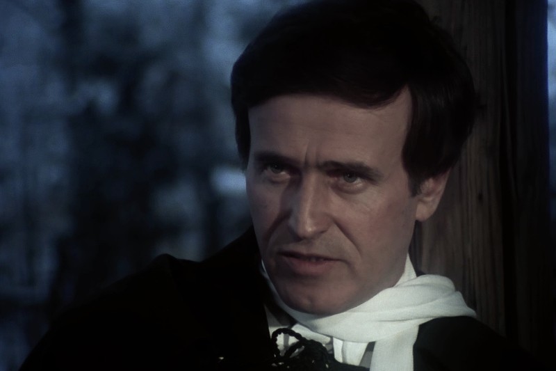 Mężczyzna o ciemnych włosach i poważnym wyrazie twarzy, ubrany w czarny płaszcz i biały szalik, stoi przed drewnianym słupem na tle rozmytego pleneru.