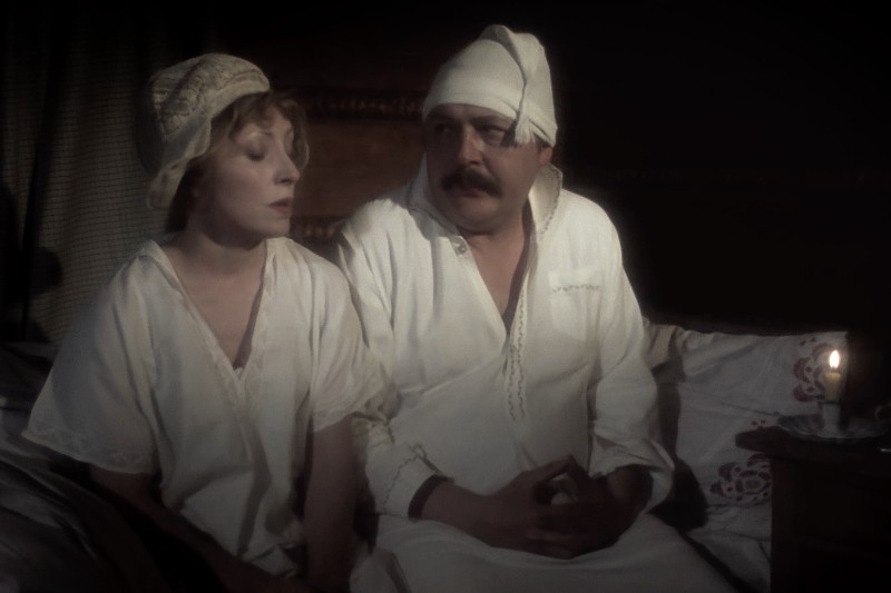 Mężczyzna i kobieta siedzą na łóżku ubrani w staromodne piżamy i czepki nocne, oświetleni małą świecą na stoliku nocnym. Pokój wydaje się ciemny, a oboje wyglądają poważnie lub zamyśleni.