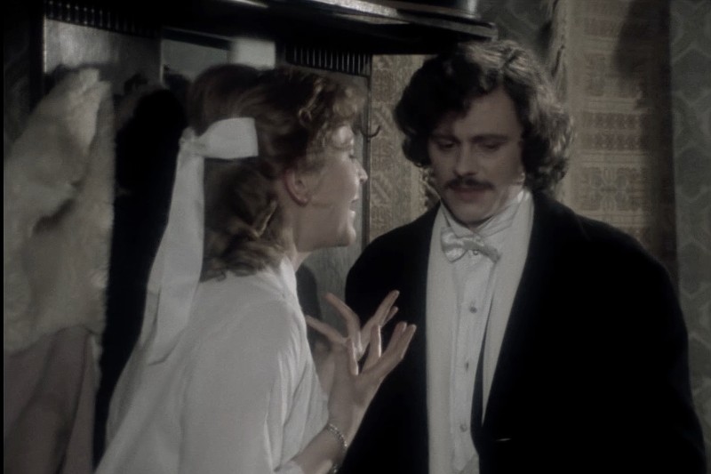 Kobieta w białej bluzce ze wstążką we włosach gestykuluje namiętnie, rozmawiając z mężczyzną o kręconych włosach i wąsach, ubranym formalnie w czarną marynarkę i białą muszkę, w pomieszczeniu w stylu vintage.