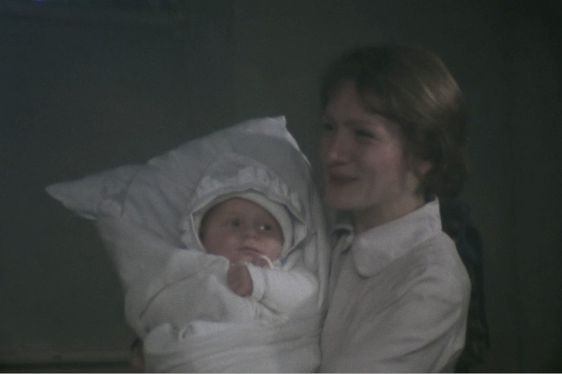Uśmiechnięta kobieta trzymająca dziecko owinięte w biały kocyk i czepek. Tło jest słabo oświetlone, co tworzy delikatną, intymną atmosferę.