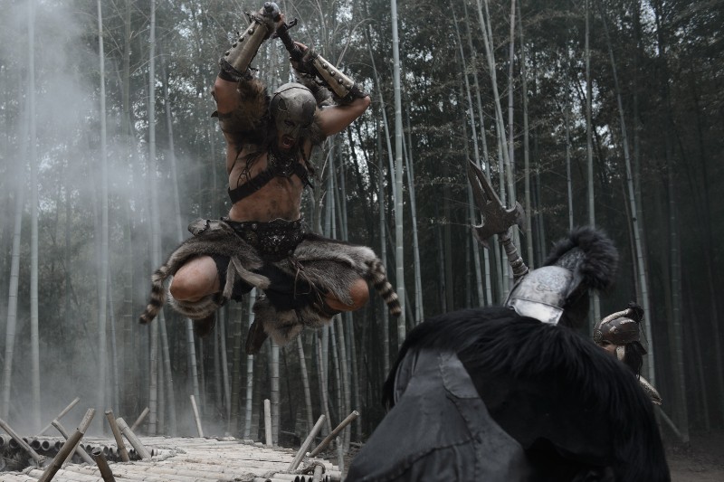 Zaciekły wojownik w futrze i zbroi skacze w powietrzu z podniesioną bronią, szykując się do ataku na opancerzonego przeciwnika w bambusowym lesie. Mgła i dramatyczne oświetlenie wzmacniają intensywną scenę bitwy.