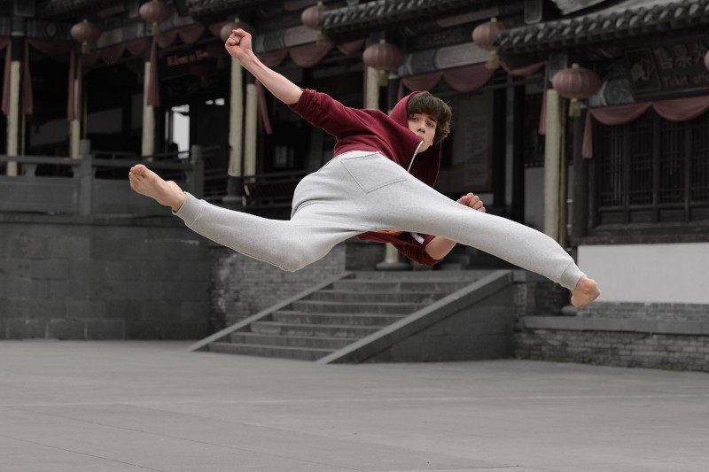Osoba ubrana w bordową bluzę z kapturem i szare spodnie dresowe wykonuje wysoki kopnięcie w stylu sztuk walki na dziedzińcu na świeżym powietrzu, w tle widać tradycyjną azjatycką architekturę.