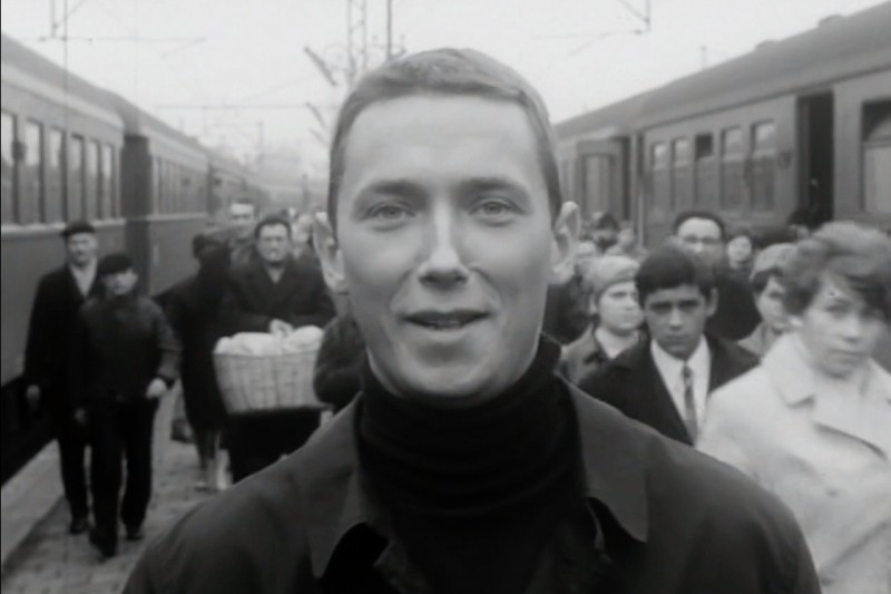 Młody mężczyzna w golfie stoi i uśmiecha się na pierwszym planie na peronie kolejowym, za nim widać tłum ludzi i dwa pociągi w czerni i bieli.