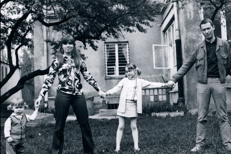 Stare czarno-białe zdjęcie czteroosobowej rodziny stojącej na podwórku, trzymającej się za ręce. Dwoje dorosłych i dwoje dzieci stoją w szeregu przed domem z otwartymi oknami i drzewami w pobliżu.