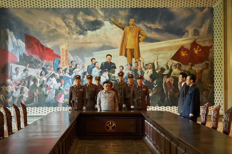 Grupa umundurowanych oficerów i urzędników stoi przy długim stole przed dużym muralem przedstawiającym przywódcę, czerwone flagi i wiwatujące tłumy. Pomieszczenie ma ozdobne ściany, a puste krzesła ustawione są wzdłuż stołu.