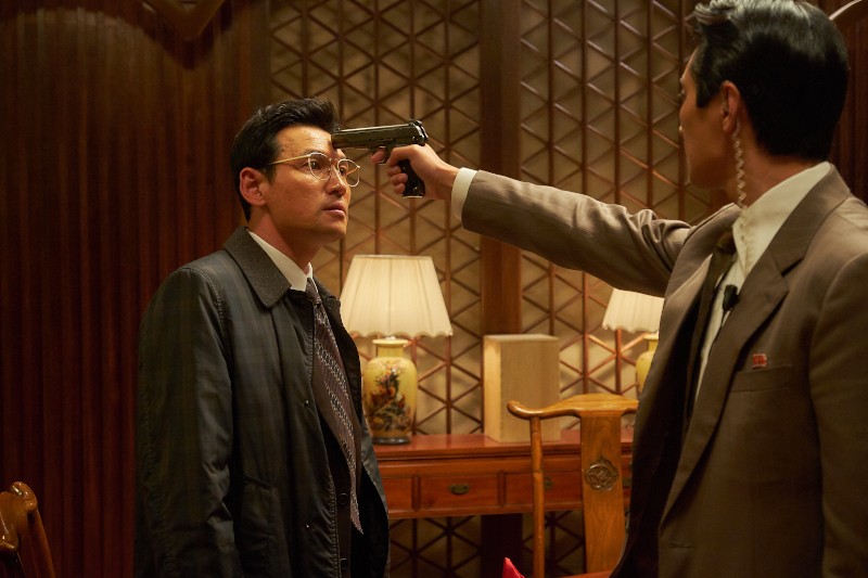 Mężczyzna w okularach stoi spokojnie, podczas gdy inny mężczyzna w garniturze przykłada mu pistolet do głowy w słabo oświetlonym, wyłożonym drewnem pokoju z ozdobną lampą i składanym parawanem w tle.