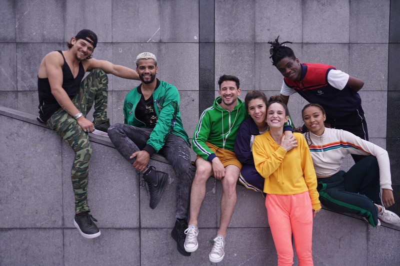 Siedmiu młodych dorosłych ubranych w codzienne, kolorowe stroje siedzi i opiera się o szarą kamienną ścianę, uśmiecha się i pozują szczęśliwie w miejskim otoczeniu.