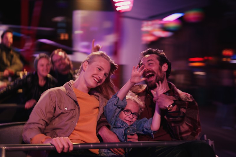 Radosna rodzina składająca się z trzech osób — dwóch dorosłych i dziecka — uśmiecha się i śmieje podczas jazdy na karuzeli w parku rozrywki, w tle widać kolorowe światła i rozmazane postacie.