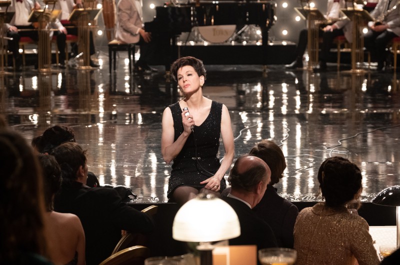 Kobieta w czarnej sukience siedzi na skraju sceny, trzyma mikrofon i śpiewa przed publicznością w słabo oświetlonym, eleganckim pomieszczeniu z pianinem i muzykami w tle.