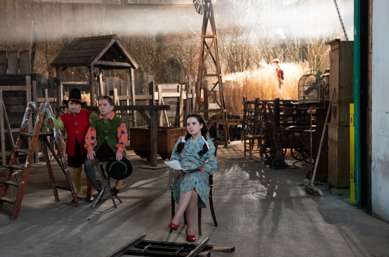 Trzy osoby w kolorowych kostiumach siedzą w rustykalnym, zagraconym zestawie z drewnianymi rekwizytami i sprzętem rolniczym. Jedna kobieta w niebieskim wzorzystym płaszczu siedzi z przodu, trzymając notatnik, podczas gdy światło spływa z góry.