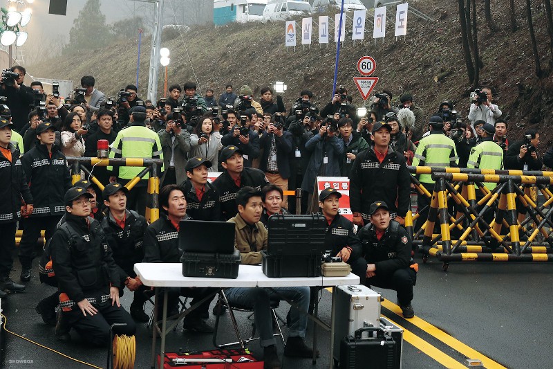 Grupa umundurowanych ratowników i funkcjonariuszy siedzi i stoi wokół sprzętu na drodze, podczas gdy duży tłum fotografów i gapiów zbiera się za żółtymi barykadami.