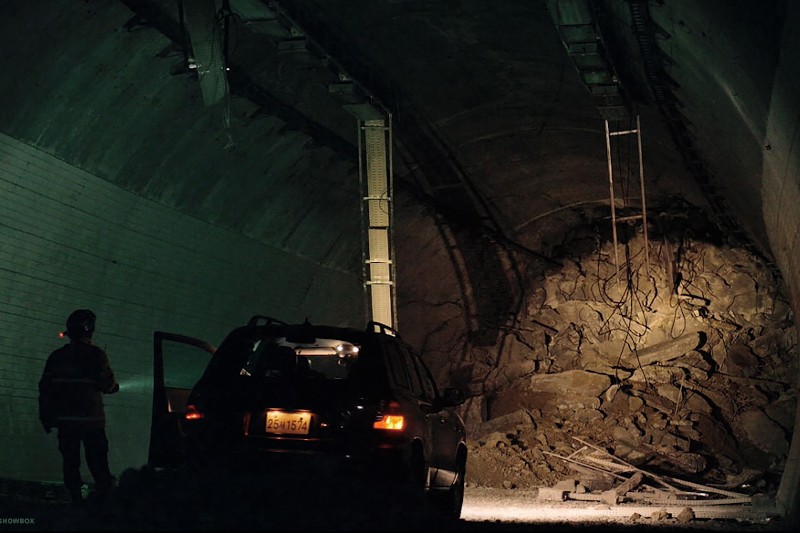 Samochód z otwartymi drzwiami stoi w słabo oświetlonym tunelu, naprzeciwko dużej sterty opadłych kamieni i gruzu blokującego przejazd. Osoba stoi obok samochodu, patrząc w stronę gruzu.