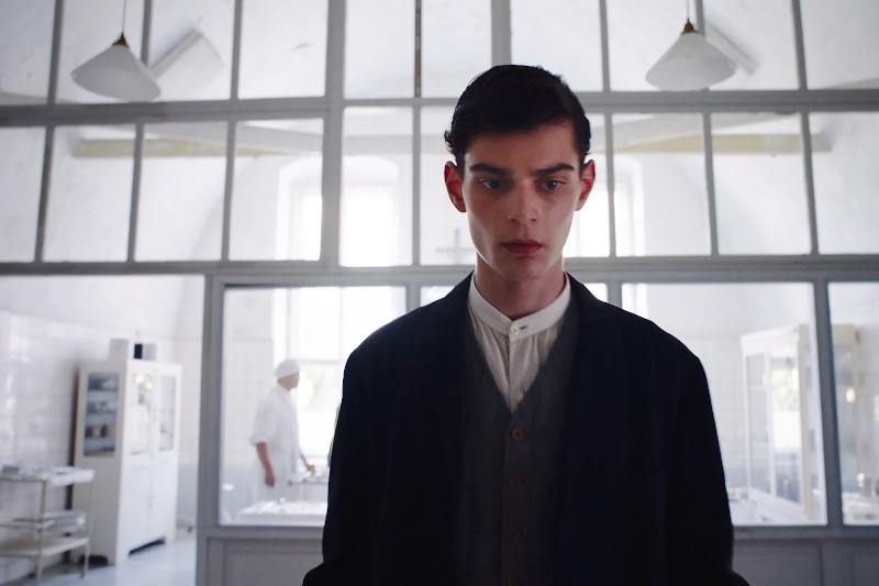 Młody mężczyzna o ciemnych włosach i formalnym ubraniu stoi w jasnym, klinicznym pomieszczeniu ze szklanymi ścianami; za nim w tle rozmazuje się postać ubrana na biało, prawdopodobnie kucharz lub pracownik służby zdrowia.