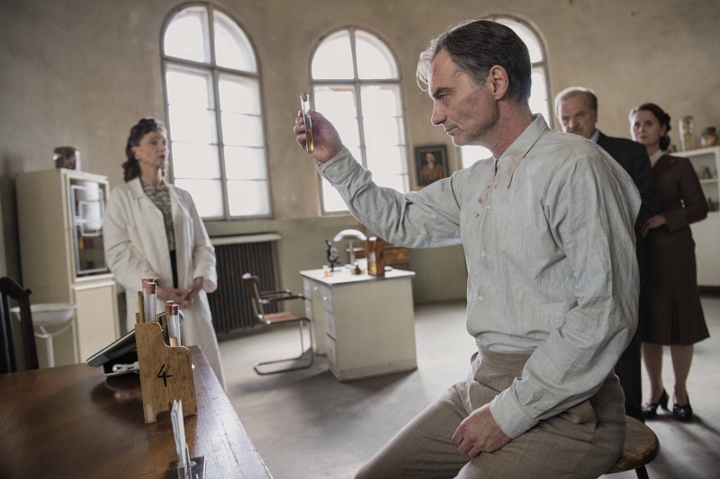 Mężczyzna w ubraniu vintage bada probówkę w laboratorium, a inne osoby, w tym kobieta w fartuchu laboratoryjnym, obserwują go. Pomieszczenie ma duże okna łukowe i zawiera sprzęt naukowy.