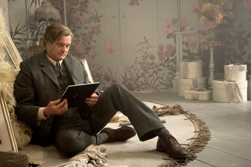 Mężczyzna w szarym garniturze siedzi na podłodze, opierając się o krzesło i zamyślony patrząc na oprawione zdjęcie. W pokoju jest tapeta w kwiaty, pudła na kapelusze i delikatne oświetlenie, tworząc atmosferę vintage.