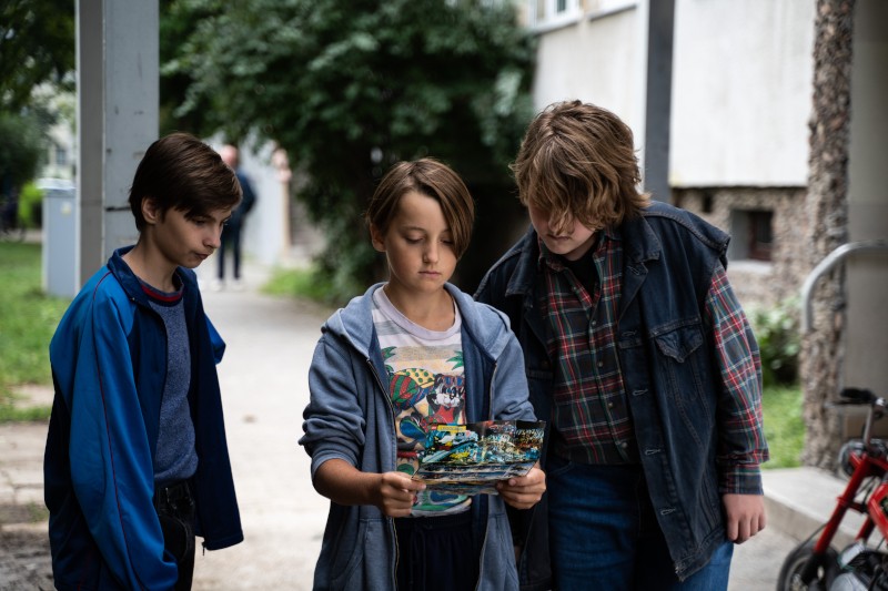 Trzech chłopców stoi na zewnątrz, w pobliżu budynku. Chłopiec w środku trzyma i patrzy na kolorową mapę lub magazyn, podczas gdy dwaj chłopcy obok niego patrzą ciekawie. W tle widać drzewa i trawę.