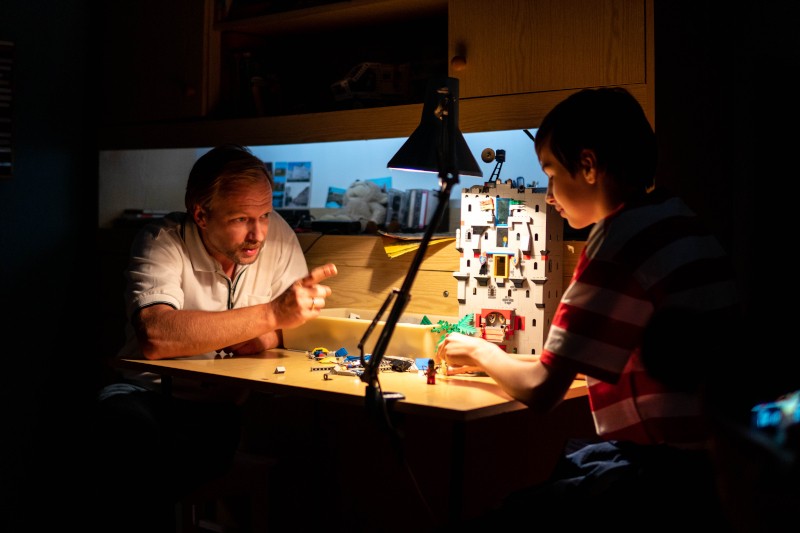 Mężczyzna i chłopiec siedzą przy biurku pod lampą, budując z klocków LEGO. Mężczyzna gestykuluje ożywiony, podczas gdy chłopiec słucha, obaj skupieni na swojej twórczej aktywności w przytulnym, słabo oświetlonym pokoju.
