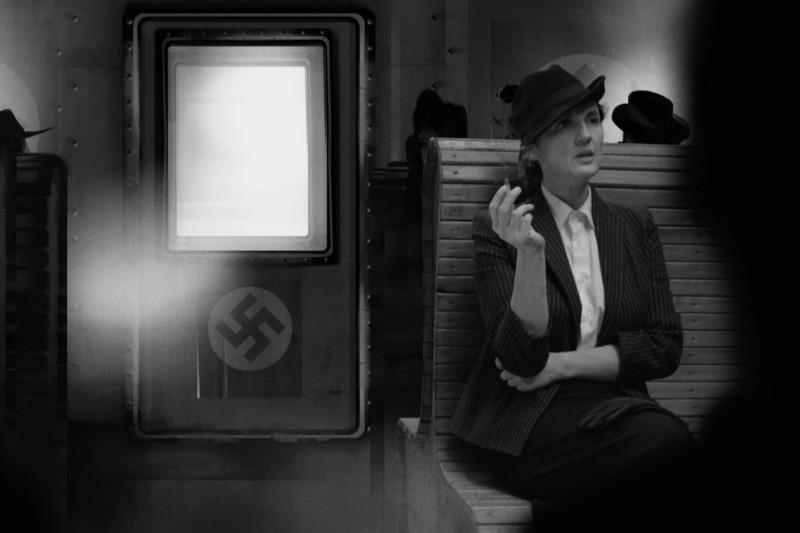 Kobieta w garniturze i kapeluszu siedzi na ławce, trzymając fajkę. Za nią stoi maszyna z dużym symbolem swastyki i jasnym, pustym ekranem. Obraz jest czarno-biały.