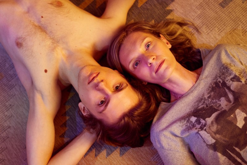 Mężczyzna bez koszuli i kobieta w nadrukowanym T-shircie leżą obok siebie na wzorzystej powierzchni, patrząc w górę na kamerę z neutralnymi wyrazami twarzy. Ich głowy są blisko siebie, a łagodne oświetlenie tworzy ciepłą atmosferę.