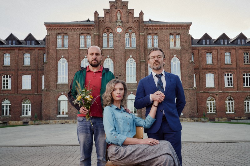 Trzech dorosłych pozuje przed dużym ceglanym budynkiem. Kobieta siedzi, a dwóch mężczyzn stoi obok niej; jeden trzyma bukiet, a drugi trzyma ją za rękę. Wszyscy mają poważne miny.