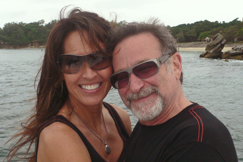 Uśmiechnięta para w okularach przeciwsłonecznych obejmuje się nad wodą, w tle widać drzewa i skały wzdłuż linii brzegowej.