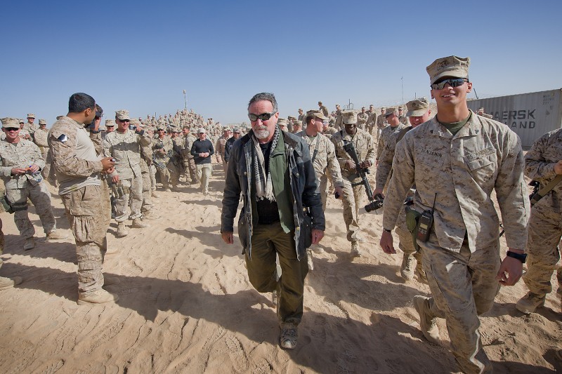 Mężczyzna w cywilnym ubraniu idzie po piaszczystym gruncie otoczony przez dużą grupę umundurowanych żołnierzy pod czystym, błękitnym niebem. Niektórzy żołnierze wchodzą w interakcję, podczas gdy inni patrzą w stronę kamery.