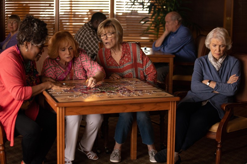 Cztery starsze kobiety siedzą przy stole, pracując nad układanką, podczas gdy inna starsza kobieta siedzi sama z boku, wyglądając na zamyśloną. Inne osoby są widoczne w tle ciepło oświetlonego pokoju.
