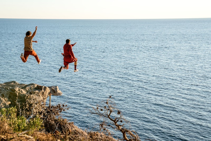 Dwie osoby skaczą ze skalistego klifu w stronę oceanu poniżej w słoneczny dzień, a rozległa błękitna woda rozciąga się aż po horyzont. Na pierwszym planie widać kilka krzaków.
