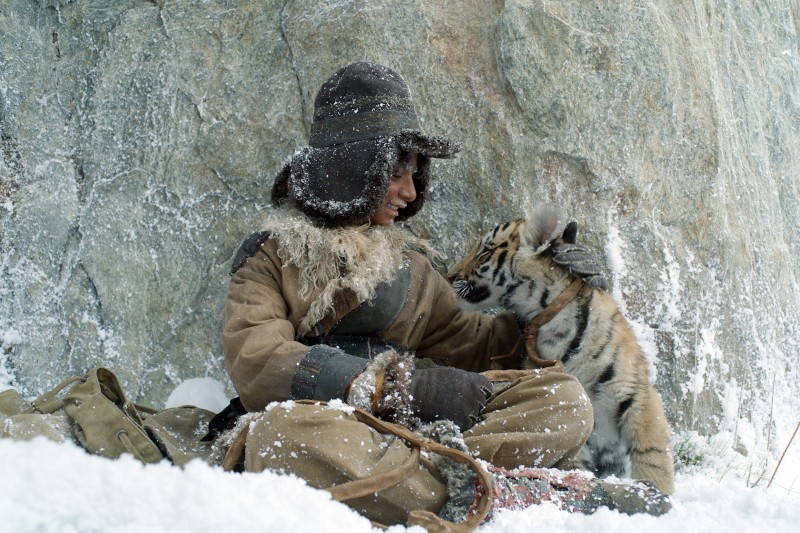 Osoba ubrana w grube zimowe ubranie siedzi na śniegu, uśmiecha się i rozmawia z tygryskiem, który znajduje się obok niej na tle ośnieżonych skał.