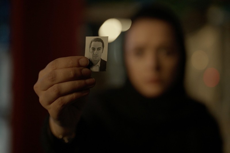 Osoba na ciemnym, rozmytym tle trzyma w stronę aparatu małe, czarno-białe zdjęcie twarzy mężczyzny, na którym widać wyraźnie jego dłoń i zdjęcie.