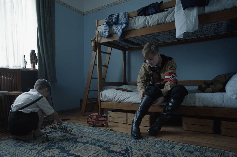 Dwóch chłopców w słabo oświetlonym pokoju z piętrowymi łóżkami; jeden nosi mundur Hitlerjugend i wysokie, czarne buty, podczas gdy drugi siedzi na podłodze i bawi się żołnierzykami i czerwoną ciężarówką.