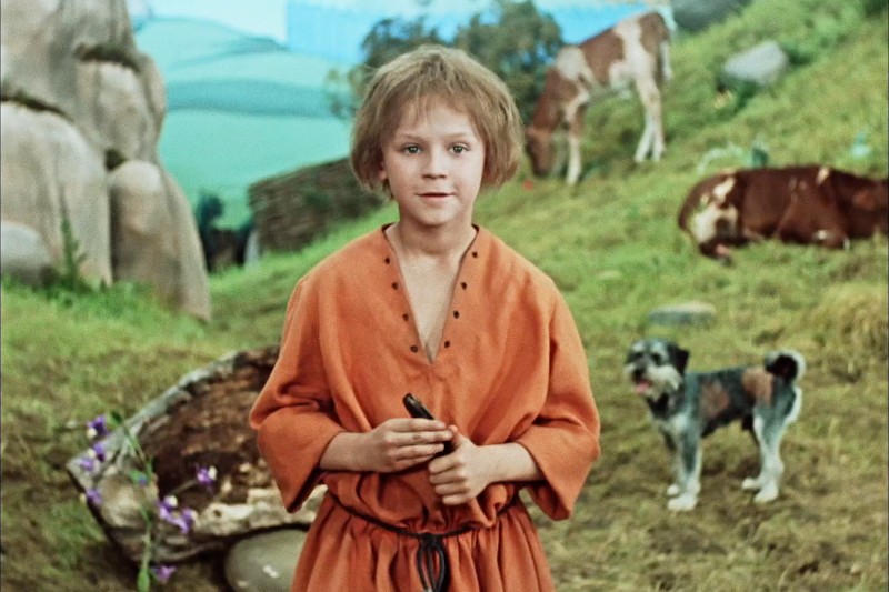 Małe dziecko w pomarańczowej tunice stoi trzymając mały przedmiot, a w tle widać psa i krowy pasące się na zielonej trawie i skałach. Scena wydaje się sielska i spokojna.